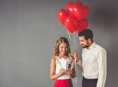 A man holds a bunch of balloons for his girlfriend as she reads his card, while they work on celebrating Valentine's Day sober